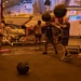 Military personnel are conducting a fitness workout session in the hangar bay of a naval aircraft carrier. Sailors are performing medicine ball exercises in a group setting, with a parked military aircraft (numbered 402) and fitness equipment visible in the background. A 'Beaver Fit' branded fitness station and a whiteboard with workout instructions are visible to the right.