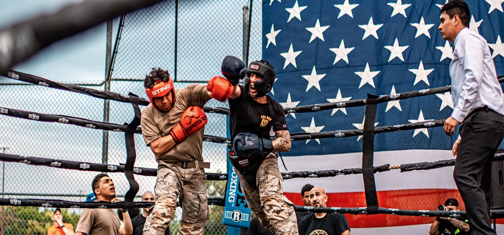 Two fighters in military camouflage uniforms are competing in an outdoor boxing match inside a professional ring. A large American flag is prominently displayed in the background, suggesting this is a military-organized sporting event. A referee in a white dress shirt observes the action from ringside.