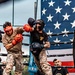 Two fighters in military camouflage uniforms are competing in an outdoor boxing match inside a professional ring. A large American flag is prominently displayed in the background, suggesting this is a military-organized sporting event. A referee in a white dress shirt observes the action from ringside.
