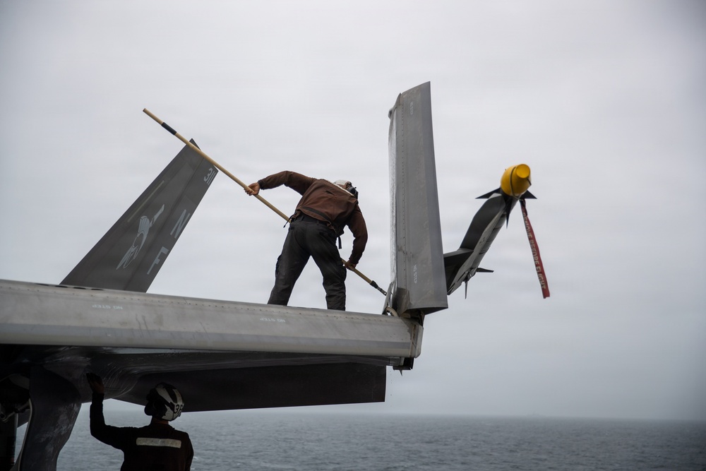 A maintenance technician in a brown jacket is standing on the wing of a military jet aircraft, using a long-handled tool to clean or inspect the surface. The aircraft appears to be on the deck of an aircraft carrier over open ocean. A yellow safety cap and red remove-before-flight streamer are visible on what appears to be a missile or weapon mounted on the aircraft.