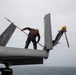 A maintenance technician in a brown jacket is standing on the wing of a military jet aircraft, using a long-handled tool to clean or inspect the surface. The aircraft appears to be on the deck of an aircraft carrier over open ocean. A yellow safety cap and red remove-before-flight streamer are visible on what appears to be a missile or weapon mounted on the aircraft.