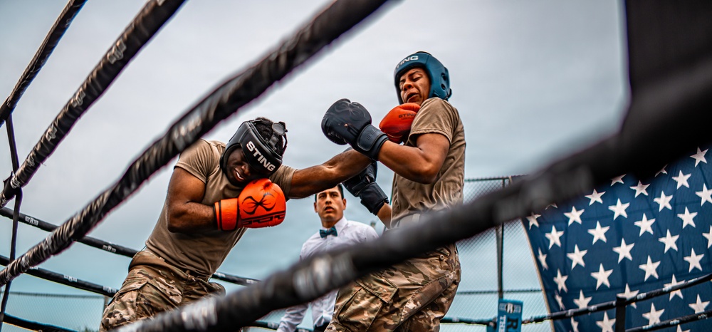 Two individuals in military camouflage uniforms are engaged in an outdoor boxing match inside a ring. One boxer wearing orange gloves and a black STING headgear is exchanging punches with an opponent wearing blue gloves and a teal STING headgear. A referee in a white shirt and bow tie oversees the bout, with an American flag visible in the background.