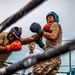 Two individuals in military camouflage uniforms are engaged in an outdoor boxing match inside a ring. One boxer wearing orange gloves and a black STING headgear is exchanging punches with an opponent wearing blue gloves and a teal STING headgear. A referee in a white shirt and bow tie oversees the bout, with an American flag visible in the background.