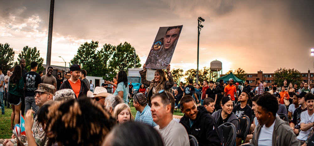 A large outdoor gathering at dusk features a diverse crowd of people, with a woman prominently holding up a large portrait photograph of a person in military uniform. The event appears to be a military-related celebration or homecoming, suggested by the military portrait, camouflage clothing visible on some attendees, and the festive atmosphere. The scene is set against a dramatic sunset sky with storm clouds, trees, a water tower, and what appears to be a military or institutional campus in the background.