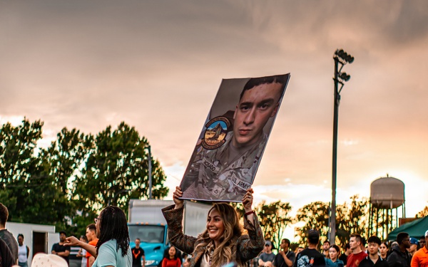 A large outdoor gathering at dusk features a diverse crowd of people, with a woman prominently holding up a large portrait photograph of a person in military uniform. The event appears to be a military-related celebration or homecoming, suggested by the military portrait, camouflage clothing visible on some attendees, and the festive atmosphere. The scene is set against a dramatic sunset sky with storm clouds, trees, a water tower, and what appears to be a military or institutional campus in the background.