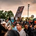 A large outdoor gathering at dusk features a diverse crowd of people, with a woman prominently holding up a large portrait photograph of a person in military uniform. The event appears to be a military-related celebration or homecoming, suggested by the military portrait, camouflage clothing visible on some attendees, and the festive atmosphere. The scene is set against a dramatic sunset sky with storm clouds, trees, a water tower, and what appears to be a military or institutional campus in the background.