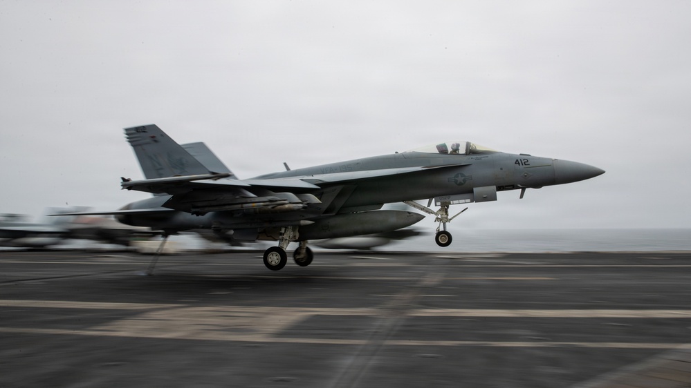 A U.S. Navy F/A-18 Super Hornet (bureau number 412) is captured launching or landing on an aircraft carrier flight deck. The aircraft has its landing gear deployed and appears to be in a low-altitude, high-speed maneuver over the carrier deck. The overcast sky and ocean horizon are visible in the background.