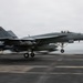 A U.S. Navy F/A-18 Super Hornet (bureau number 412) is captured launching or landing on an aircraft carrier flight deck. The aircraft has its landing gear deployed and appears to be in a low-altitude, high-speed maneuver over the carrier deck. The overcast sky and ocean horizon are visible in the background.