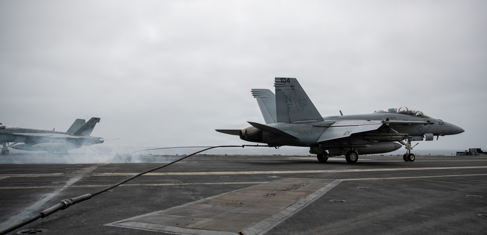 A U.S. Navy F/A-18 Super Hornet marked '104' with tail code 'NF' is positioned on an aircraft carrier flight deck, connected to an arresting wire or launch cable. In the background, another aircraft is launching or landing with visible exhaust smoke and steam. The overcast sky and carrier deck environment indicate active flight operations at sea.