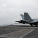 A U.S. Navy F/A-18 Super Hornet marked '104' with tail code 'NF' is positioned on an aircraft carrier flight deck, connected to an arresting wire or launch cable. In the background, another aircraft is launching or landing with visible exhaust smoke and steam. The overcast sky and carrier deck environment indicate active flight operations at sea.