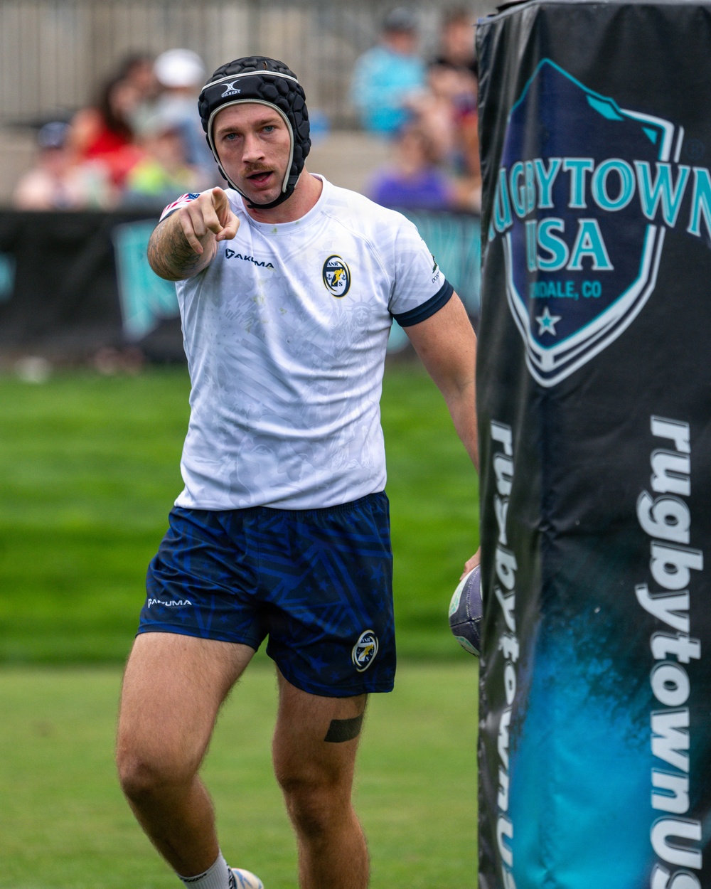 A male rugby player wearing a white Pakuma jersey, blue shorts, and a black Gilbert headguard is pointing toward the camera during a match at Rugbytown USA in Glendale, Colorado. He is holding a rugby ball near a branded banner post and appears to be communicating with a teammate or official. The background shows a green playing field and blurred spectators in the stands.
