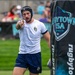 A male rugby player wearing a white Pakuma jersey, blue shorts, and a black Gilbert headguard is pointing toward the camera during a match at Rugbytown USA in Glendale, Colorado. He is holding a rugby ball near a branded banner post and appears to be communicating with a teammate or official. The background shows a green playing field and blurred spectators in the stands.