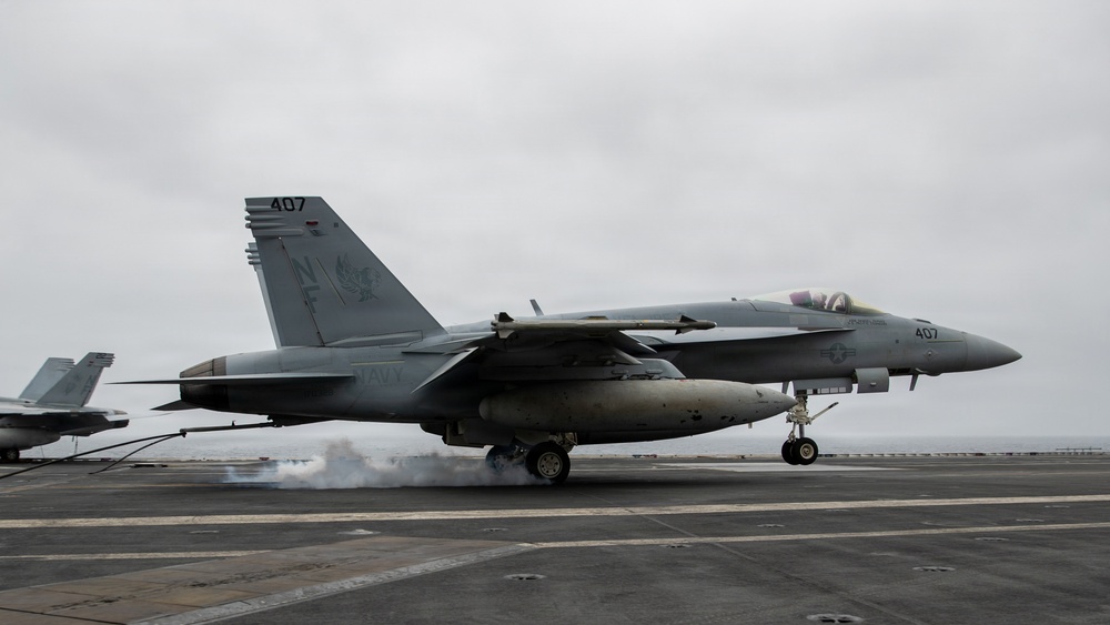 A U.S. Navy F/A-18 Super Hornet (Bureau Number 407) is captured landing on an aircraft carrier deck, with visible tire smoke from touchdown. The aircraft bears the tail code 'NF' and squadron markings, indicating it belongs to a carrier air wing based in the Western Pacific. The overcast sky and carrier deck environment suggest active flight operations at sea.