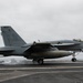 A U.S. Navy F/A-18 Super Hornet (Bureau Number 407) is captured landing on an aircraft carrier deck, with visible tire smoke from touchdown. The aircraft bears the tail code 'NF' and squadron markings, indicating it belongs to a carrier air wing based in the Western Pacific. The overcast sky and carrier deck environment suggest active flight operations at sea.
