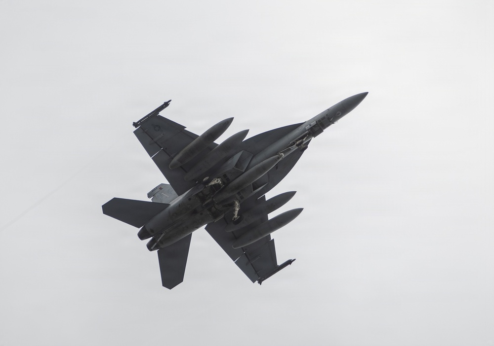 A dark-colored military jet fighter is photographed from below against an overcast gray sky. The aircraft appears to be in a banking maneuver, revealing its underside with multiple external stores mounted on wing pylons. The twin-tail configuration and overall design are clearly visible.