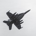 A dark-colored military jet fighter is photographed from below against an overcast gray sky. The aircraft appears to be in a banking maneuver, revealing its underside with multiple external stores mounted on wing pylons. The twin-tail configuration and overall design are clearly visible.