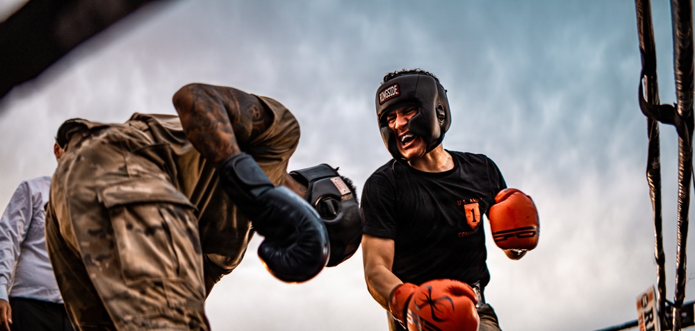 Two individuals are engaged in an outdoor boxing match inside a ring. The fighter on the right wears a black 'Ringside' headguard, red boxing gloves, and a black t-shirt with 'U.S. Army' text visible. The fighter on the left wears camouflage clothing and black boxing gloves, delivering or blocking a punch.