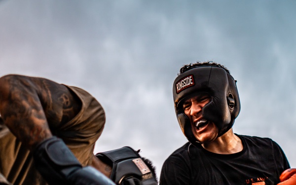 Two individuals are engaged in an outdoor boxing match inside a ring. The fighter on the right wears a black 'Ringside' headguard, red boxing gloves, and a black t-shirt with 'U.S. Army' text visible. The fighter on the left wears camouflage clothing and black boxing gloves, delivering or blocking a punch.