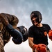 Two individuals are engaged in an outdoor boxing match inside a ring. The fighter on the right wears a black 'Ringside' headguard, red boxing gloves, and a black t-shirt with 'U.S. Army' text visible. The fighter on the left wears camouflage clothing and black boxing gloves, delivering or blocking a punch.