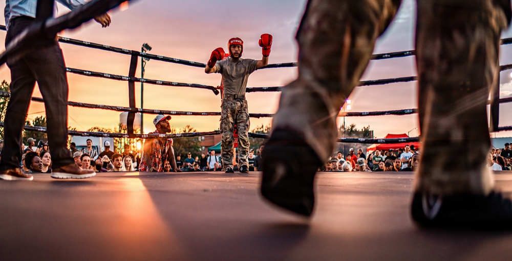 An outdoor boxing match is taking place at sunset, with a fighter in camouflage pants and red boxing gloves standing in the center of the ring with arms raised. The low-angle shot is taken from ring level, capturing the legs of another fighter or referee in the foreground. A large crowd of spectators surrounds the ring, watching the event under a dramatic pink and orange sky.