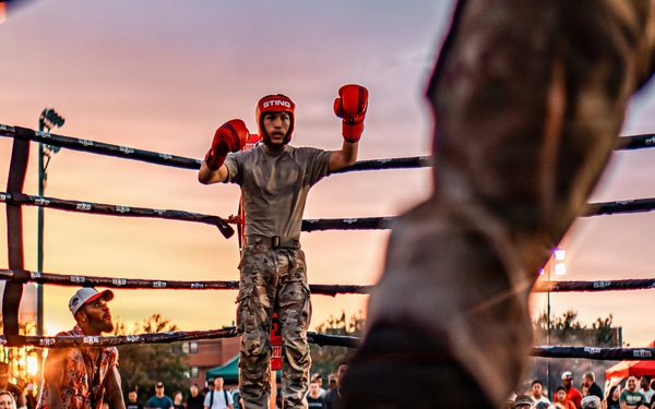 An outdoor boxing match is taking place at sunset, with a fighter in camouflage pants and red boxing gloves standing in the center of the ring with arms raised. The low-angle shot is taken from ring level, capturing the legs of another fighter or referee in the foreground. A large crowd of spectators surrounds the ring, watching the event under a dramatic pink and orange sky.