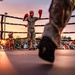 An outdoor boxing match is taking place at sunset, with a fighter in camouflage pants and red boxing gloves standing in the center of the ring with arms raised. The low-angle shot is taken from ring level, capturing the legs of another fighter or referee in the foreground. A large crowd of spectators surrounds the ring, watching the event under a dramatic pink and orange sky.