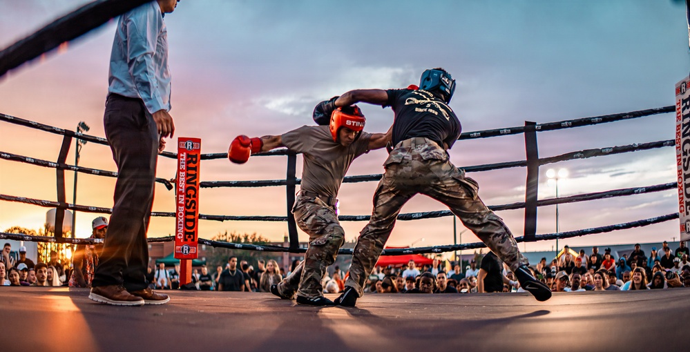 Two fighters wearing military camouflage uniforms and boxing gear are competing in an outdoor boxing ring at dusk. A referee in a white shirt and dark pants observes the bout closely. The event appears to be a military boxing competition held before a large crowd.