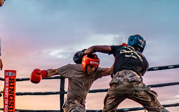 Two fighters wearing military camouflage uniforms and boxing gear are competing in an outdoor boxing ring at dusk. A referee in a white shirt and dark pants observes the bout closely. The event appears to be a military boxing competition held before a large crowd.