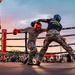 Two fighters wearing military camouflage uniforms and boxing gear are competing in an outdoor boxing ring at dusk. A referee in a white shirt and dark pants observes the bout closely. The event appears to be a military boxing competition held before a large crowd.