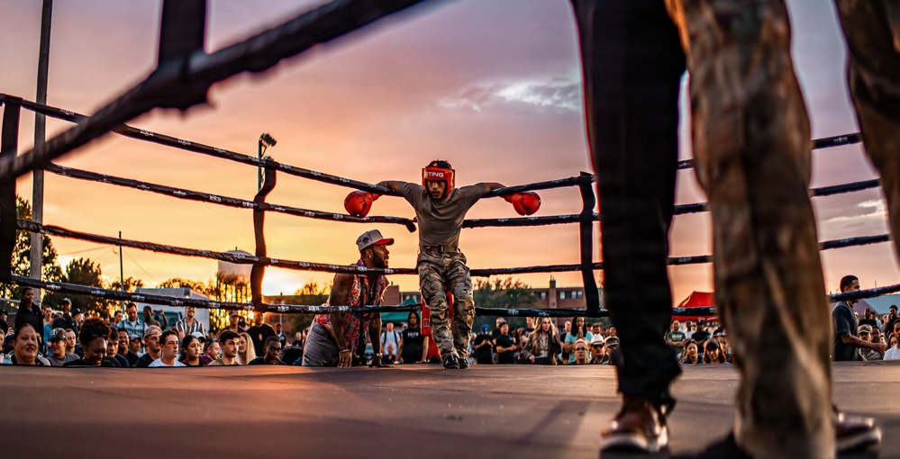 An outdoor boxing event is taking place at dusk, with a boxer in red headgear and red gloves leaning against the ring ropes in a corner. A trainer or cornerman is crouching nearby, likely providing instruction or support between rounds. A large crowd of spectators surrounds the ring, watching the event under a dramatic sunset sky.