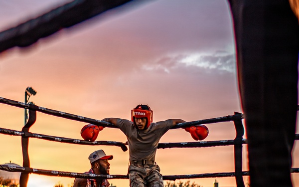 An outdoor boxing event is taking place at dusk, with a boxer in red headgear and red gloves leaning against the ring ropes in a corner. A trainer or cornerman is crouching nearby, likely providing instruction or support between rounds. A large crowd of spectators surrounds the ring, watching the event under a dramatic sunset sky.
