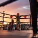 An outdoor boxing event is taking place at dusk, with a boxer in red headgear and red gloves leaning against the ring ropes in a corner. A trainer or cornerman is crouching nearby, likely providing instruction or support between rounds. A large crowd of spectators surrounds the ring, watching the event under a dramatic sunset sky.