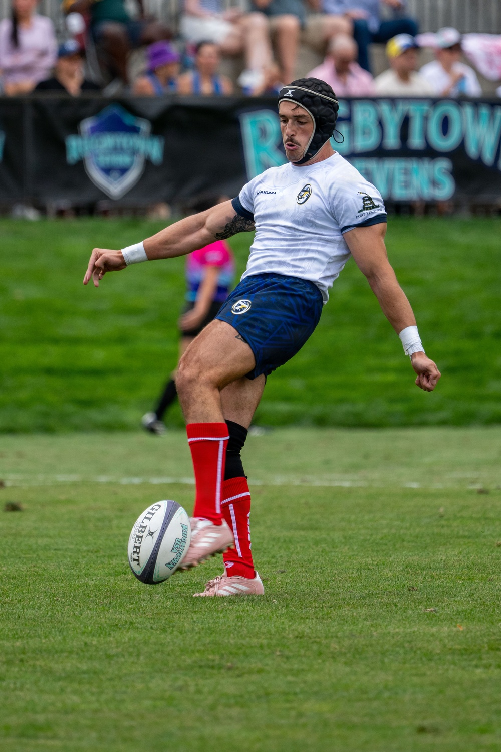 A rugby player is captured mid-kick during a match, wearing a white Yakima jersey, blue shorts, red socks, and pink cleats, along with a black protective headgear. The Gilbert rugby ball is positioned just in front of his kicking foot on a well-maintained grass field. The event appears to be 'Rugbytown Sevens' based on the visible banner signage in the background.