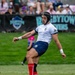 A rugby player is captured mid-kick during a match, wearing a white Yakima jersey, blue shorts, red socks, and pink cleats, along with a black protective headgear. The Gilbert rugby ball is positioned just in front of his kicking foot on a well-maintained grass field. The event appears to be 'Rugbytown Sevens' based on the visible banner signage in the background.