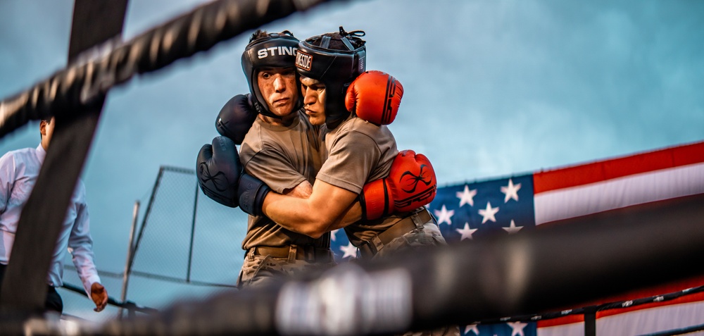 Two boxers are clinching in a boxing ring, both wearing protective headgear and gloves. One fighter wears blue gloves and a 'Sting' branded headguard, while the other wears red gloves. An American flag is prominently displayed in the background, suggesting this may be a military or patriotic sporting event.