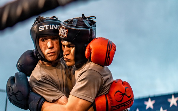 Two boxers are clinching in a boxing ring, both wearing protective headgear and gloves. One fighter wears blue gloves and a 'Sting' branded headguard, while the other wears red gloves. An American flag is prominently displayed in the background, suggesting this may be a military or patriotic sporting event.