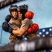 Two boxers are clinching in a boxing ring, both wearing protective headgear and gloves. One fighter wears blue gloves and a 'Sting' branded headguard, while the other wears red gloves. An American flag is prominently displayed in the background, suggesting this may be a military or patriotic sporting event.