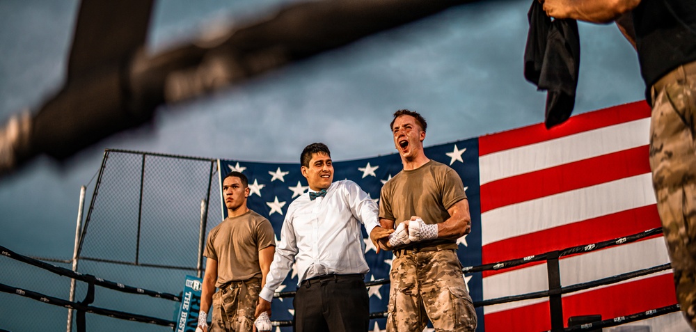 The image depicts what appears to be a military boxing event held outdoors, with a large American flag displayed prominently in the background. Three individuals are visible in the foreground near a boxing ring, two wearing military camouflage uniforms and one dressed in formal attire with a bow tie, likely serving as a referee or announcer. One of the soldiers appears to be celebrating or reacting emotionally, possibly after a bout.