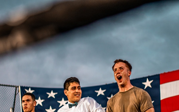 The image depicts what appears to be a military boxing event held outdoors, with a large American flag displayed prominently in the background. Three individuals are visible in the foreground near a boxing ring, two wearing military camouflage uniforms and one dressed in formal attire with a bow tie, likely serving as a referee or announcer. One of the soldiers appears to be celebrating or reacting emotionally, possibly after a bout.