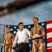 The image depicts what appears to be a military boxing event held outdoors, with a large American flag displayed prominently in the background. Three individuals are visible in the foreground near a boxing ring, two wearing military camouflage uniforms and one dressed in formal attire with a bow tie, likely serving as a referee or announcer. One of the soldiers appears to be celebrating or reacting emotionally, possibly after a bout.