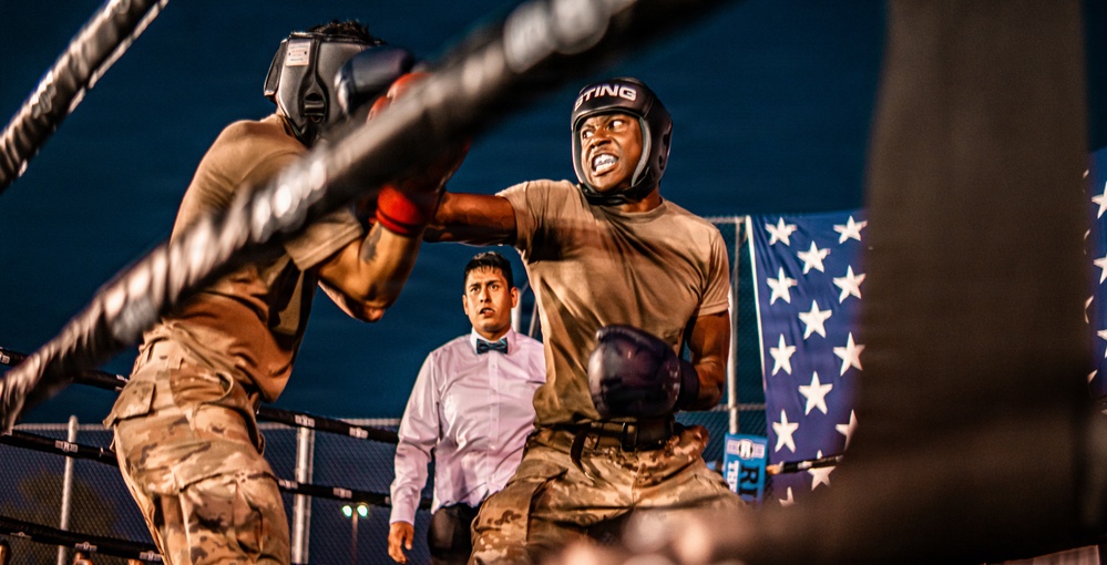 Two individuals wearing military camouflage uniforms and boxing headgear are engaged in a boxing match inside a ring. A referee in a white dress shirt and bow tie observes the bout from the background. An American flag is visible in the background, suggesting this may be a military-organized boxing event.