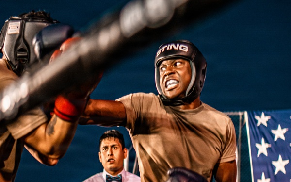 Two individuals wearing military camouflage uniforms and boxing headgear are engaged in a boxing match inside a ring. A referee in a white dress shirt and bow tie observes the bout from the background. An American flag is visible in the background, suggesting this may be a military-organized boxing event.