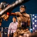 Two individuals wearing military camouflage uniforms and boxing headgear are engaged in a boxing match inside a ring. A referee in a white dress shirt and bow tie observes the bout from the background. An American flag is visible in the background, suggesting this may be a military-organized boxing event.
