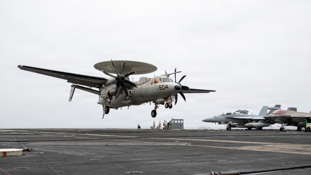 A U.S. Navy E-2 Hawkeye airborne early warning aircraft (bureau number 604) is captured on final approach for landing on an aircraft carrier flight deck. The aircraft's distinctive rotodome radar dish, twin turboprop engines, and folding wings are clearly visible as it descends with landing gear extended. Additional carrier aircraft, likely F/A-18 Hornets or Super Hornets, are parked on the starboard side of the flight deck.