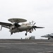 A U.S. Navy E-2 Hawkeye airborne early warning aircraft (bureau number 604) is captured on final approach for landing on an aircraft carrier flight deck. The aircraft's distinctive rotodome radar dish, twin turboprop engines, and folding wings are clearly visible as it descends with landing gear extended. Additional carrier aircraft, likely F/A-18 Hornets or Super Hornets, are parked on the starboard side of the flight deck.