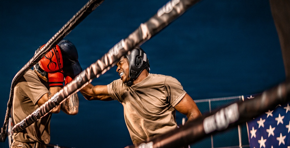 Two individuals are sparring in a boxing ring, both wearing tan/khaki t-shirts and protective headgear. The fighter on the right is delivering a punch while smiling, and the fighter on the left is wearing red boxing gloves and blocking. An American flag is partially visible in the lower right corner, suggesting a military or patriotic context.