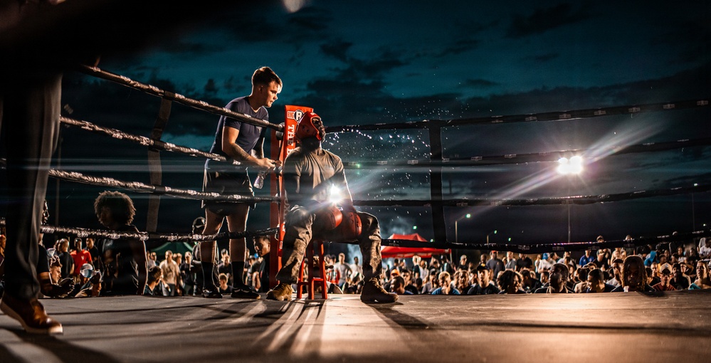 A boxing match is taking place at an outdoor night event with a large crowd in attendance. A boxer is seated on a stool in the corner of the ring during what appears to be a between-rounds break, attended by a cornerman who is providing water or treatment. The scene is dramatically lit by bright ring lights against a dark, cloudy sky.