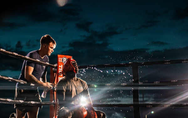 A boxing match is taking place at an outdoor night event with a large crowd in attendance. A boxer is seated on a stool in the corner of the ring during what appears to be a between-rounds break, attended by a cornerman who is providing water or treatment. The scene is dramatically lit by bright ring lights against a dark, cloudy sky.