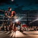 A boxing match is taking place at an outdoor night event with a large crowd in attendance. A boxer is seated on a stool in the corner of the ring during what appears to be a between-rounds break, attended by a cornerman who is providing water or treatment. The scene is dramatically lit by bright ring lights against a dark, cloudy sky.