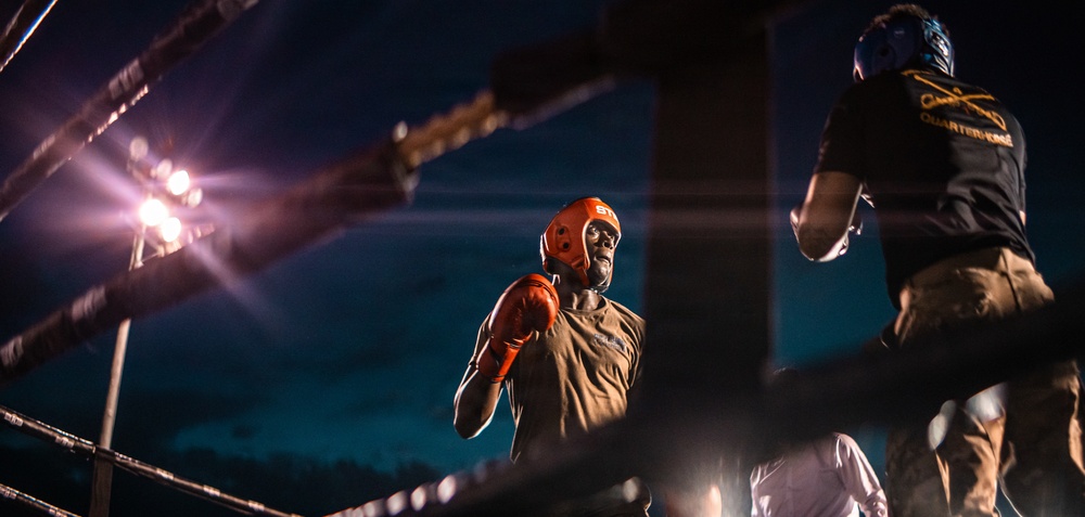 Two boxers are engaged in a match inside a boxing ring under bright artificial lights at night. The central fighter wears an orange/red protective headgear and orange boxing gloves, facing an opponent in a dark shirt with 'Quartermaster' text visible on the back. The dramatic low-angle shot and ring ropes frame the intense athletic confrontation.